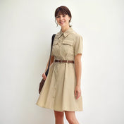 Femme souriante portant une robe saharienne traditionnelle beige, avec ceinture marron et sac à l’épaule, posant devant un mur blanc dans un style casual chic.