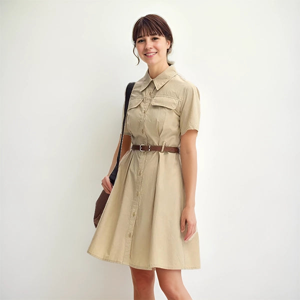 Femme souriante portant une robe saharienne traditionnelle beige, avec ceinture marron et sac à l’épaule, posant devant un mur blanc dans un style casual chic.