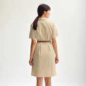 Vue arrière d’une femme portant une robe saharienne traditionnelle beige ceinturée, cheveux lâchés et posée debout devant un fond blanc.