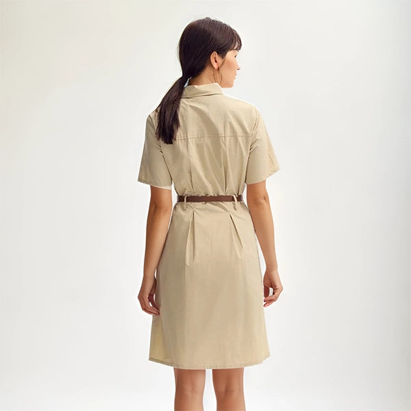 Vue arrière d’une femme portant une robe saharienne traditionnelle beige ceinturée, cheveux lâchés et posée debout devant un fond blanc.