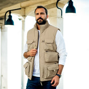 Un homme portant une veste saharienne sans manche de couleur beige, se tenant debout sous un porche éclairé par des lampes suspendues. Il est habillé d'une chemise blanche et d'un pantalon bleu, complétant son look élégant et décontracté, idéal pour les journées fraîches d'automne.