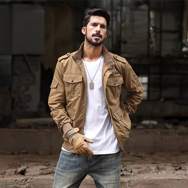Un homme portant une veste de style baroudeur en couleur marron beige, debout dans un environnement urbain délabré. Il est habillé d'un t-shirt blanc et d'un jean, complétant son look aventureux avec des gants assortis. Sa posture confiante et son regard sérieux reflètent un style robuste et pratique, parfait pour un homme actif et stylé.