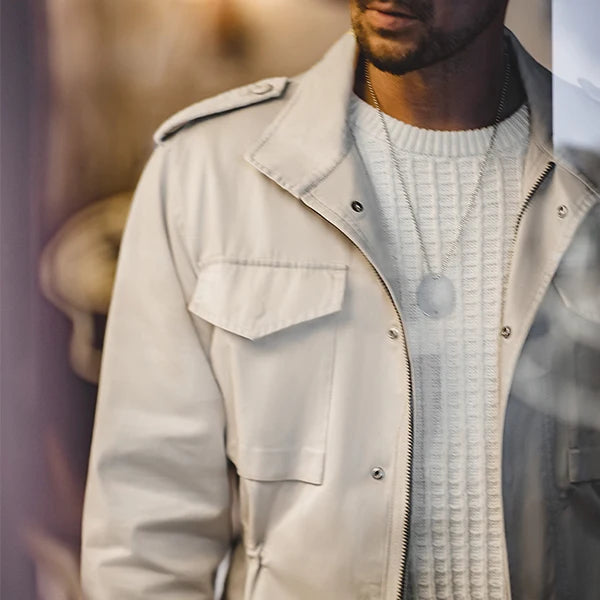 Portrait en gros plan d'un homme vêtu d'une veste saharienne blanche, caractérisée par un col large et des poches à rabat. Sous la veste, il porte un pull blanc en maille épaisse. L'ensemble crée une tenue élégante et polyvalente, idéale pour une allure sophistiquée en milieu urbain.