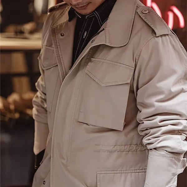 Un homme porte une veste de style saharienne en tissu beige lisse, mettant en avant un col montant, plusieurs poches plaquées sur le devant et des détails de couture précis. La veste est complétée par une chemise noire et une écharpe noire et blanche, offrant un look à la fois urbain et aventureux.