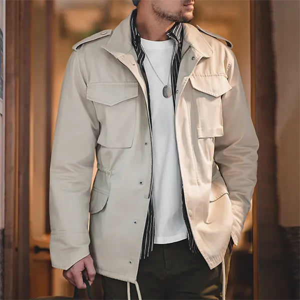 Un homme porte une veste de style saharienne de couleur beige, dotée de multiples poches plaquées et d'une ceinture ajustable à la taille. Il arbore également un foulard noir et blanc noué autour du cou, une chemise blanche légère et un pantalon sombre, complétant ainsi un look d'aventurier moderne.