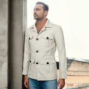 Homme portant une veste saharienne en lin de couleur claire, debout à côté d'une colonne en béton, regardant au loin. La veste est élégante avec des poches plaquées et un design à double boutonnage, parfaite pour un style décontracté chic.