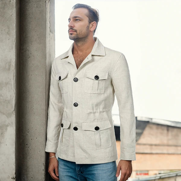 Homme portant une veste saharienne en lin de couleur claire, debout à côté d'une colonne en béton, regardant au loin. La veste est élégante avec des poches plaquées et un design à double boutonnage, parfaite pour un style décontracté chic.