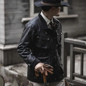 Un homme élégant portant une veste saharienne homme Barbour en noir, complétée par un chapeau assorti et tenant une canne, dégage une allure raffinée et aventurière tout en se tenant près d'un bâtiment en bois.