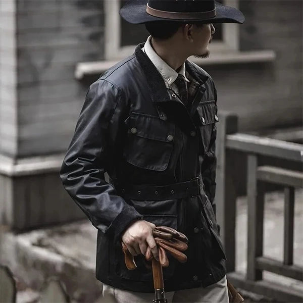 Un homme élégant portant une veste saharienne homme Barbour en noir, complétée par un chapeau assorti et tenant une canne, dégage une allure raffinée et aventurière tout en se tenant près d'un bâtiment en bois.