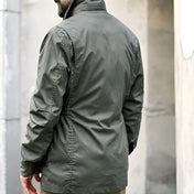 Vue arrière d'un homme portant une veste saharienne kaki, se tenant debout dans un cadre urbain. La veste est bien ajustée avec un élastique à la taille pour souligner sa silhouette. L'homme regarde au loin, ajoutant une touche de mystère à son allure générale, élégante et fonctionnelle.