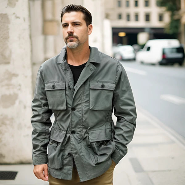 Un homme marchant confiant dans une rue urbaine, portant une veste saharienne kaki stylée. La veste comporte de multiples poches pratiques et une fermeture boutonnée, offrant un look à la fois fonctionnel et élégant. L'homme dégage une allure décontractée mais soignée, parfaitement adaptée à un environnement citadin.